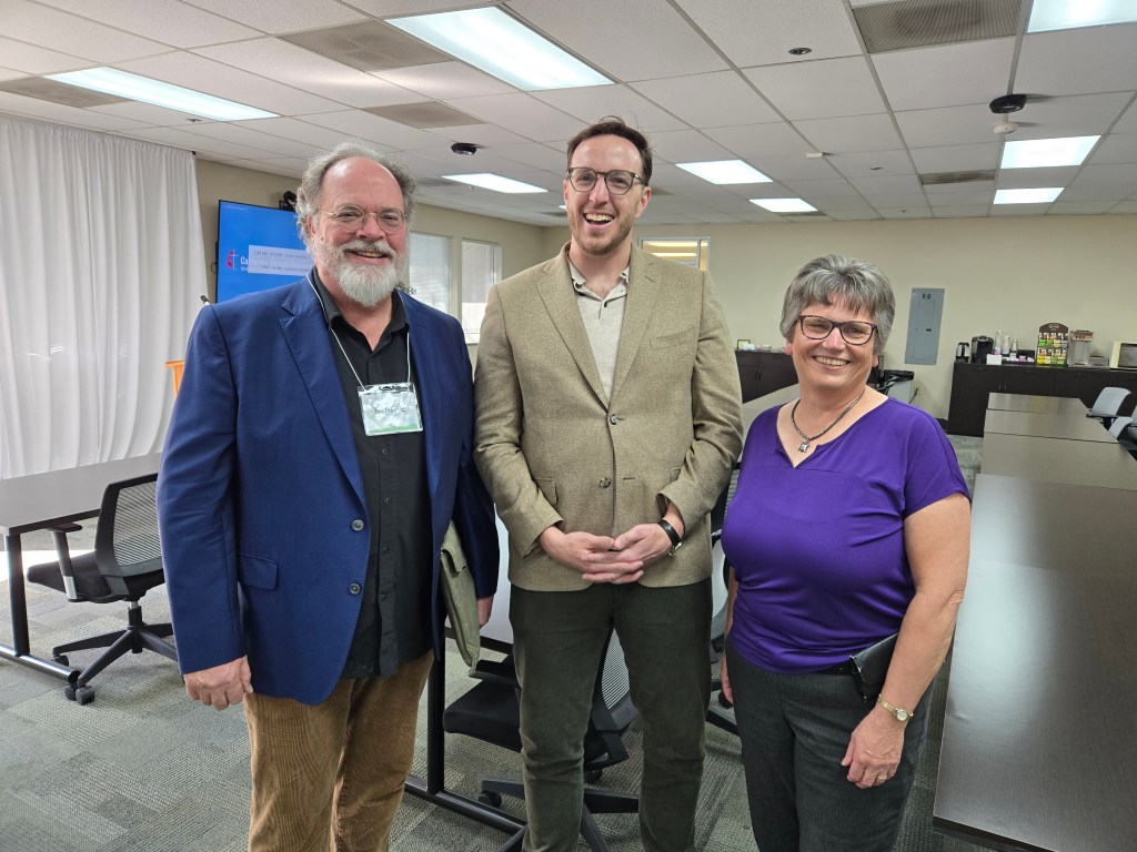 Three people standing together and smiling for a photo at the California-Nevada Conference offices in Sacramento, California. Pictured, left to right, Rev. Burke Owens, Jake Barnett, and Joan Pell.